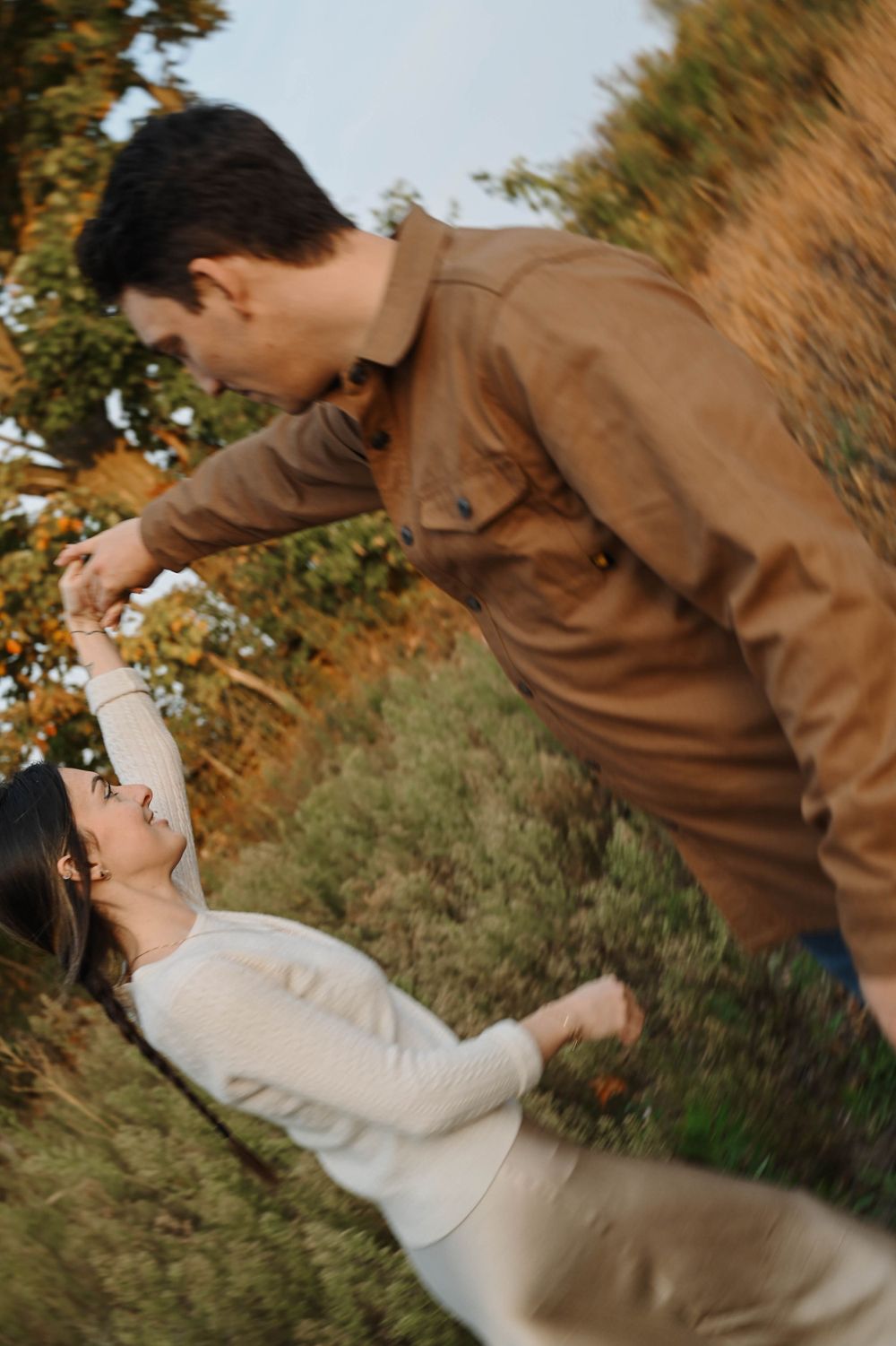 A couple in brown and white outfits playfully interact in a golden outdoor setting during sunset.