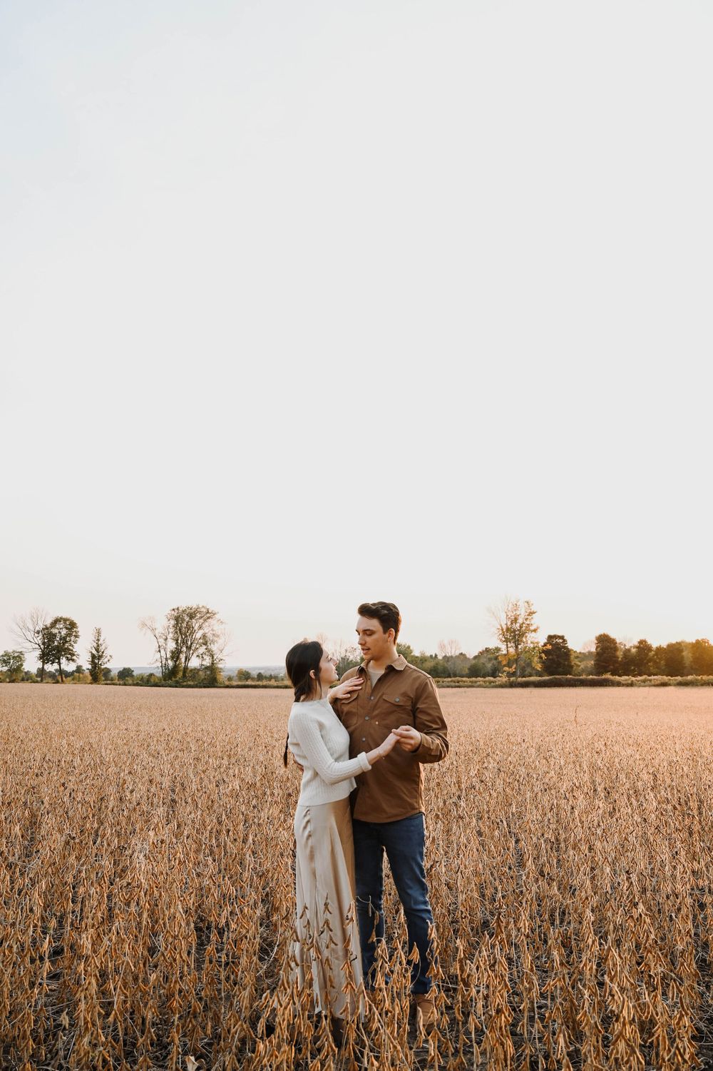 A couple stands together embracing in a golden wheat field at sunset with trees in the background.
