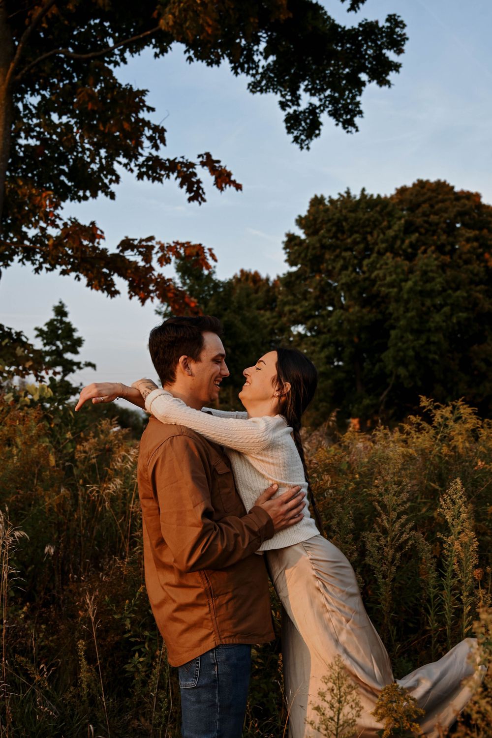 A couple embraces in a romantic moment at sunset in a field of tall grass with autumn trees in the background.