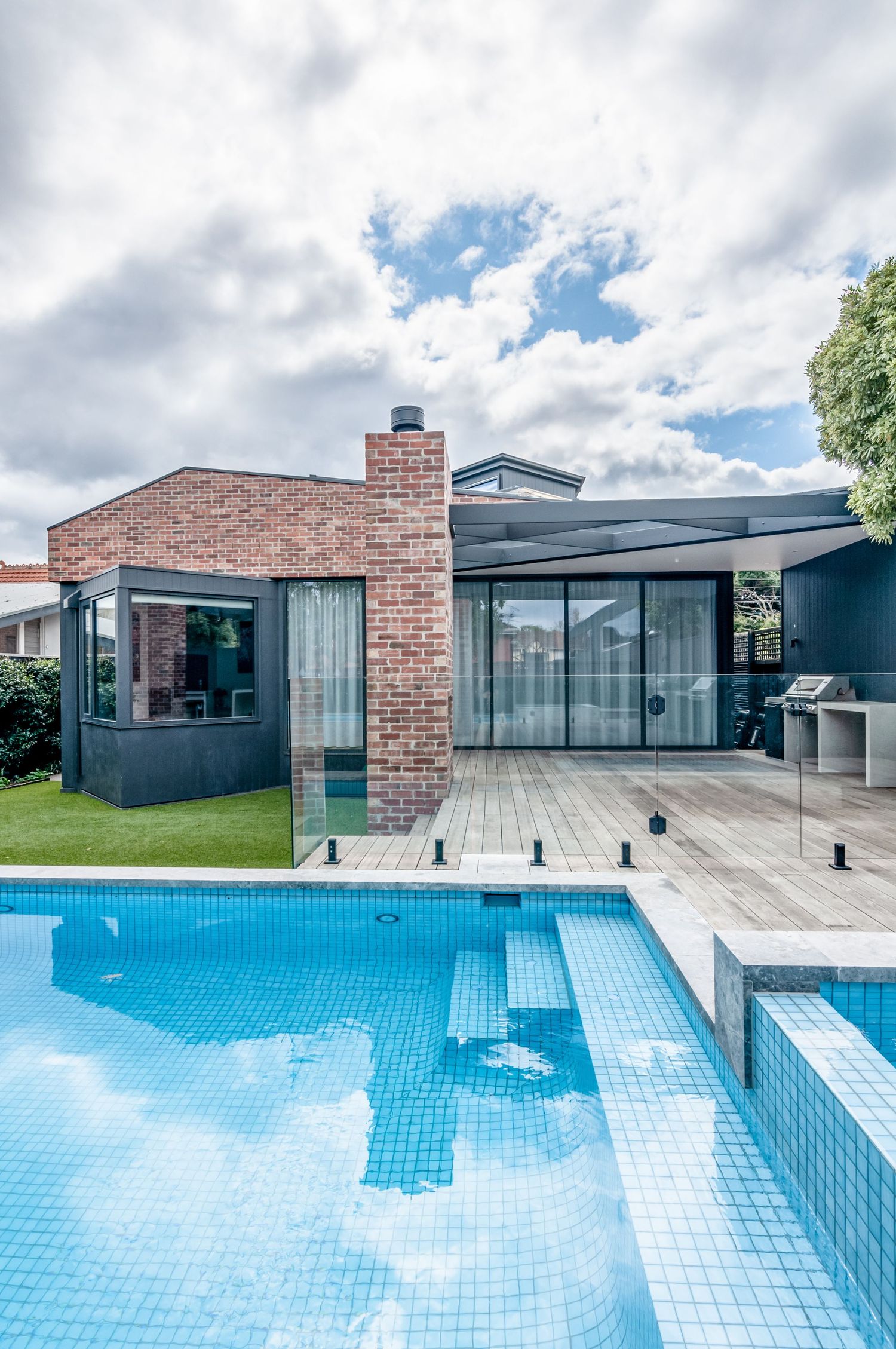 Modern house exterior with blue tiled swimming pool and glass walls under dramatic cloudy sky.