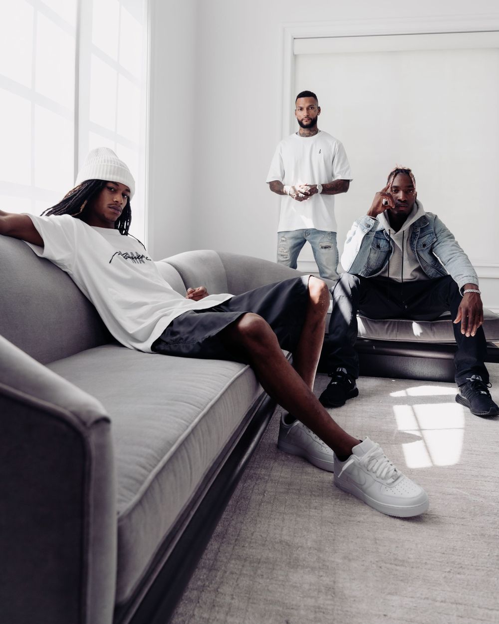 Three people relaxing in a minimalist white living room with a gray couch and casual streetwear style.
