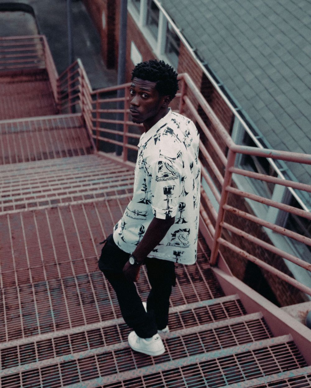 Person posing on red metal stairs wearing a patterned white button-up shirt and black pants.