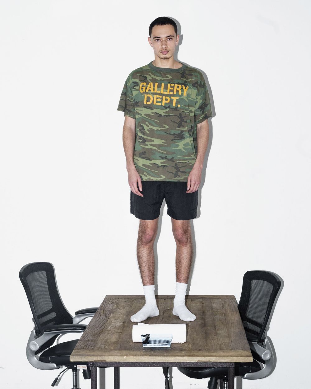 Person wearing camo Gallery Dept shirt standing on wooden table between office chairs.