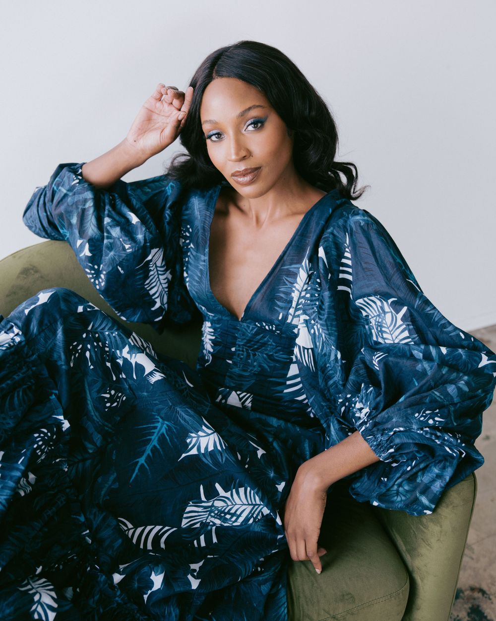 Fashion model poses in blue floral print dress while reclining on green couch.