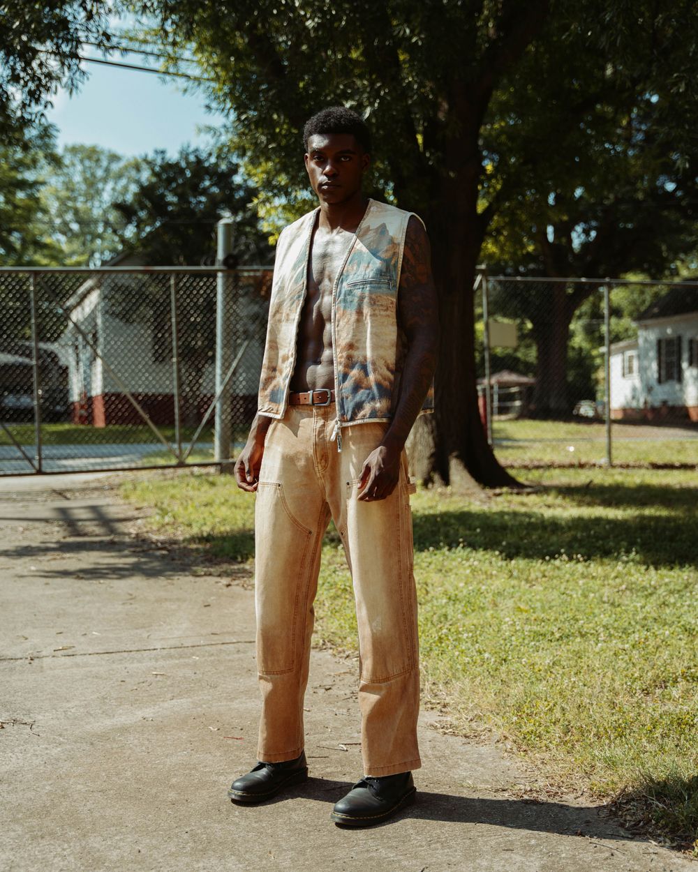 Person in light colored vest and pants standing on path with chain link fence and trees in background.