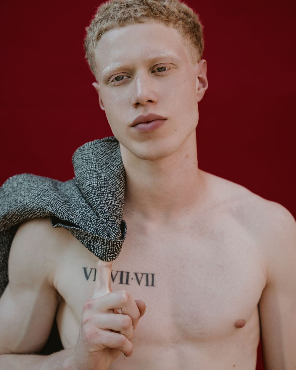 Shirtless model with roman numeral tattoo holding grey towel against red background.