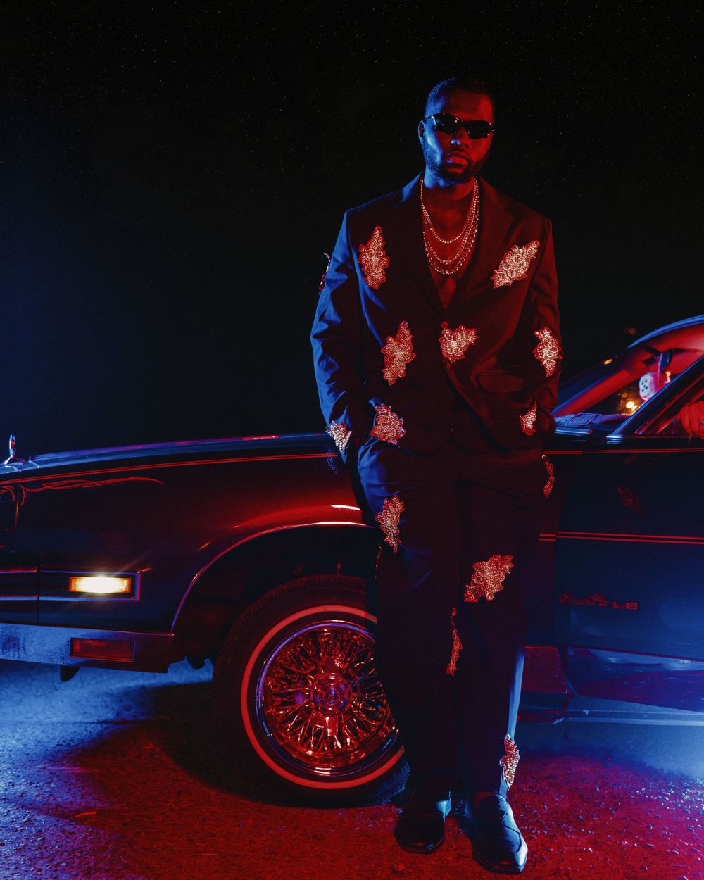 Figure in patterned jacket standing next to vintage car with neon lighting effects.