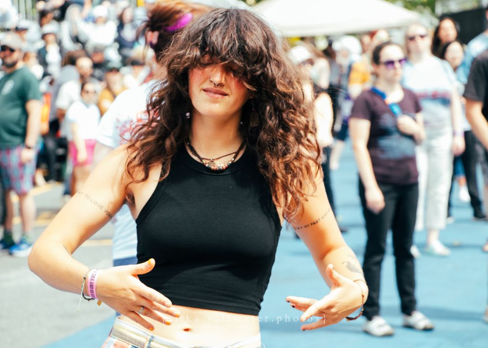A person with flowing curly brown hair dances at an outdoor event wearing a black crop top among a crowd.