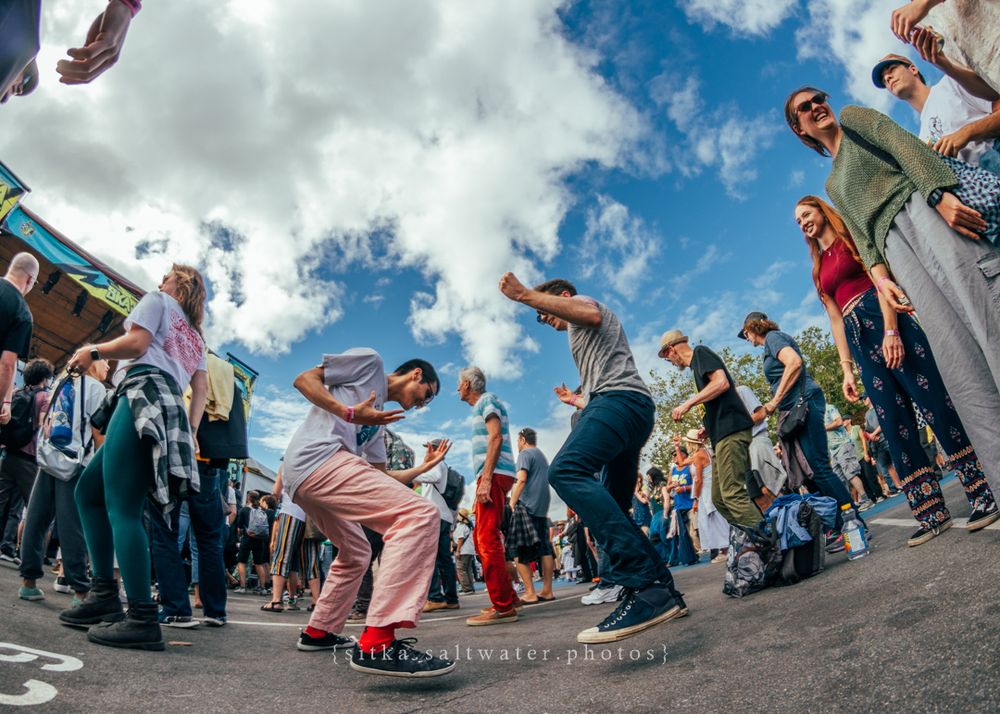 A low-angle fisheye view of street dancers performing in a circle with crowds watching under blue sky and clouds.