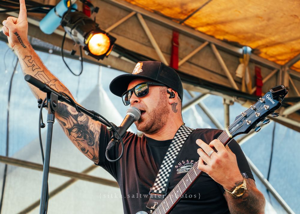 Musician wearing black sunglasses and shirt plays guitar energetically on outdoor stage during daytime performance.