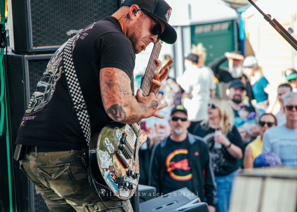 Tattooed guitarist in black t-shirt performs on stage while crowd watches during outdoor concert performance.
