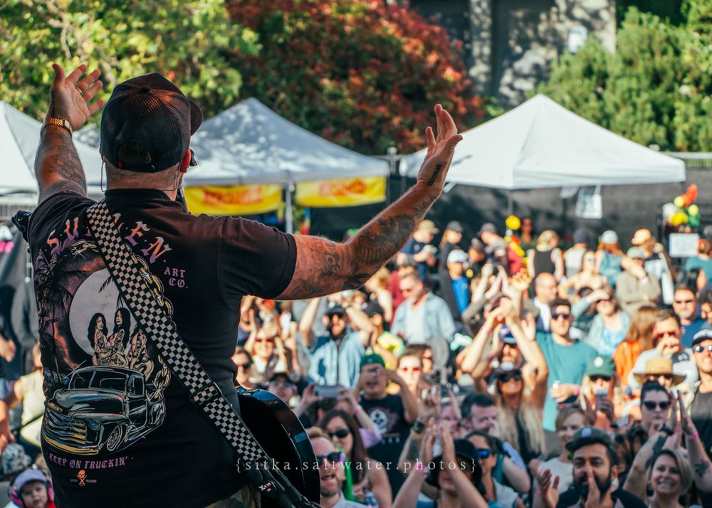 A performer on stage raises hands energetically in front of a large festival crowd with white tents in the background.