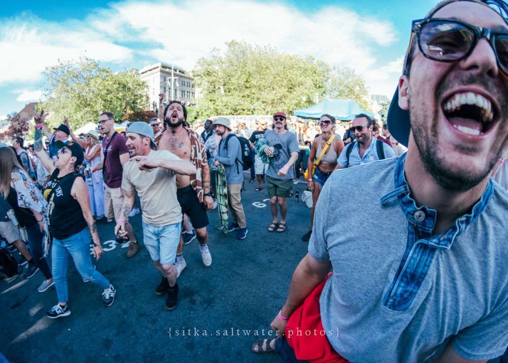 Excited festival crowd jumps and dances during an outdoor music event on a sunny day.