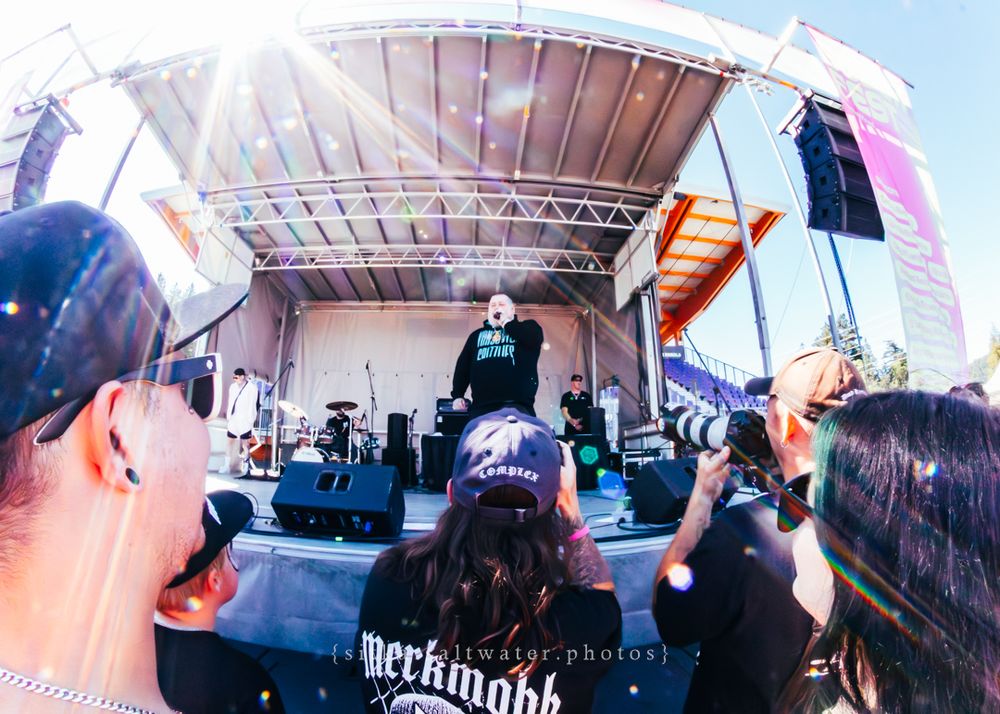 Fisheye view of a concert stage at an outdoor music festival with crowd members visible in the foreground.