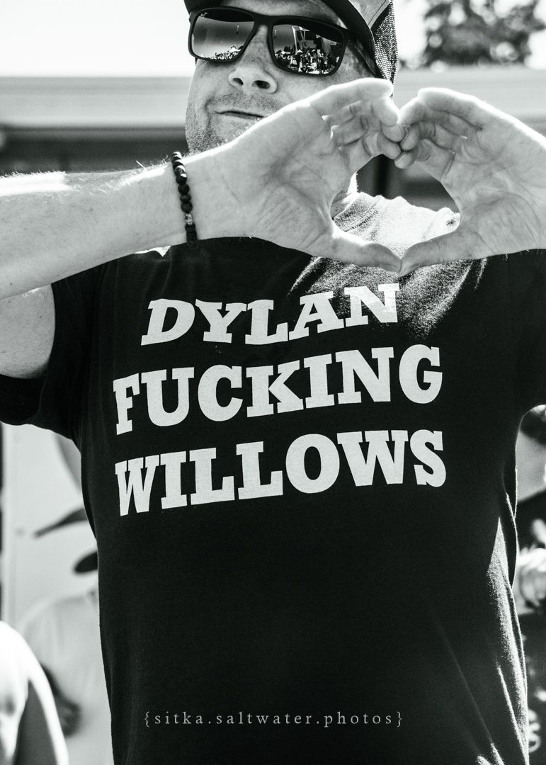 Person wearing sunglasses and black t-shirt with text 'DYLAN FUCKING WILLOWS' making heart shape gesture outdoors.