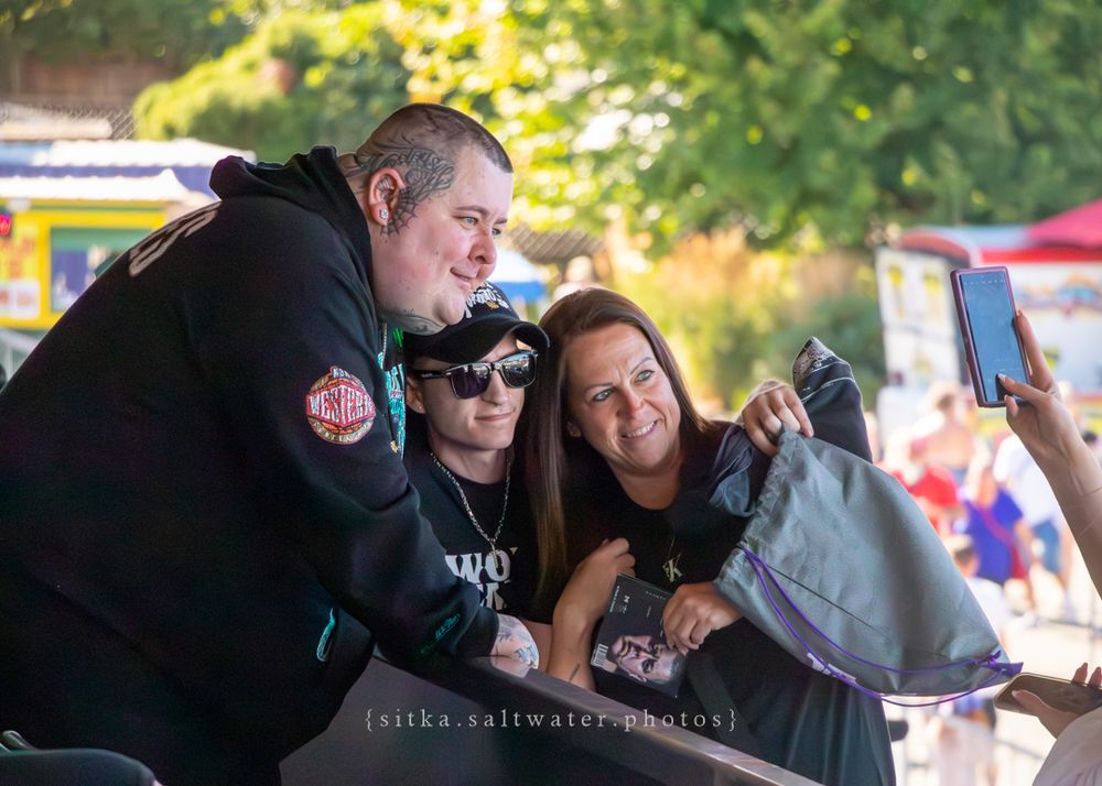Three people smile together taking a selfie during an outdoor event with blurred trees and stalls in the background.