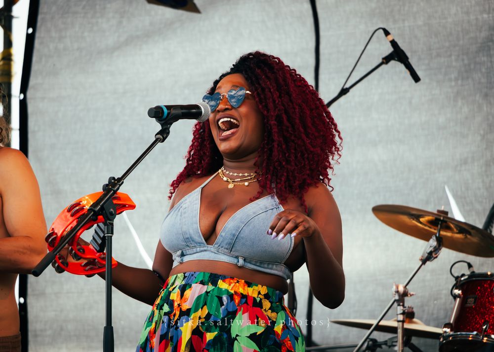 A joyful performer in a colorful skirt sings on stage with a band at an outdoor music festival.