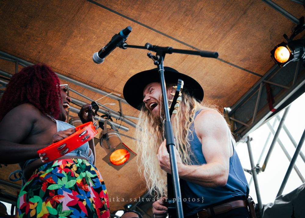 A performer with long hair and a black hat sings passionately into a microphone on stage during a live performance.