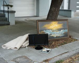 A framed seascape painting, wallet, and newspaper left on a concrete surface outdoors.