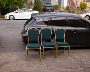 Three green banquet chairs arranged on sidewalk next to parked black car.
