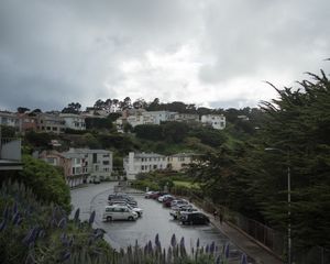 Hillside neighborhood with rows of houses and parked cars under moody overcast sky.