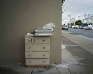 Abandoned dresser left on urban sidewalk against beige wall with city street in background.