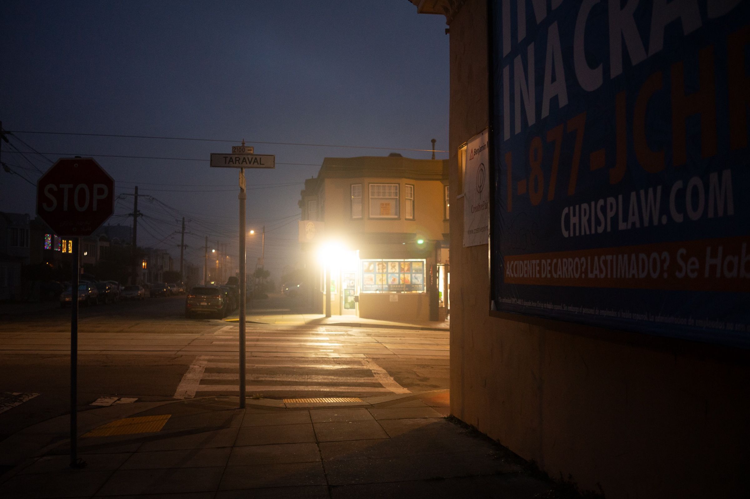 A dimly lit urban street corner at night with illuminated storefronts and signage.