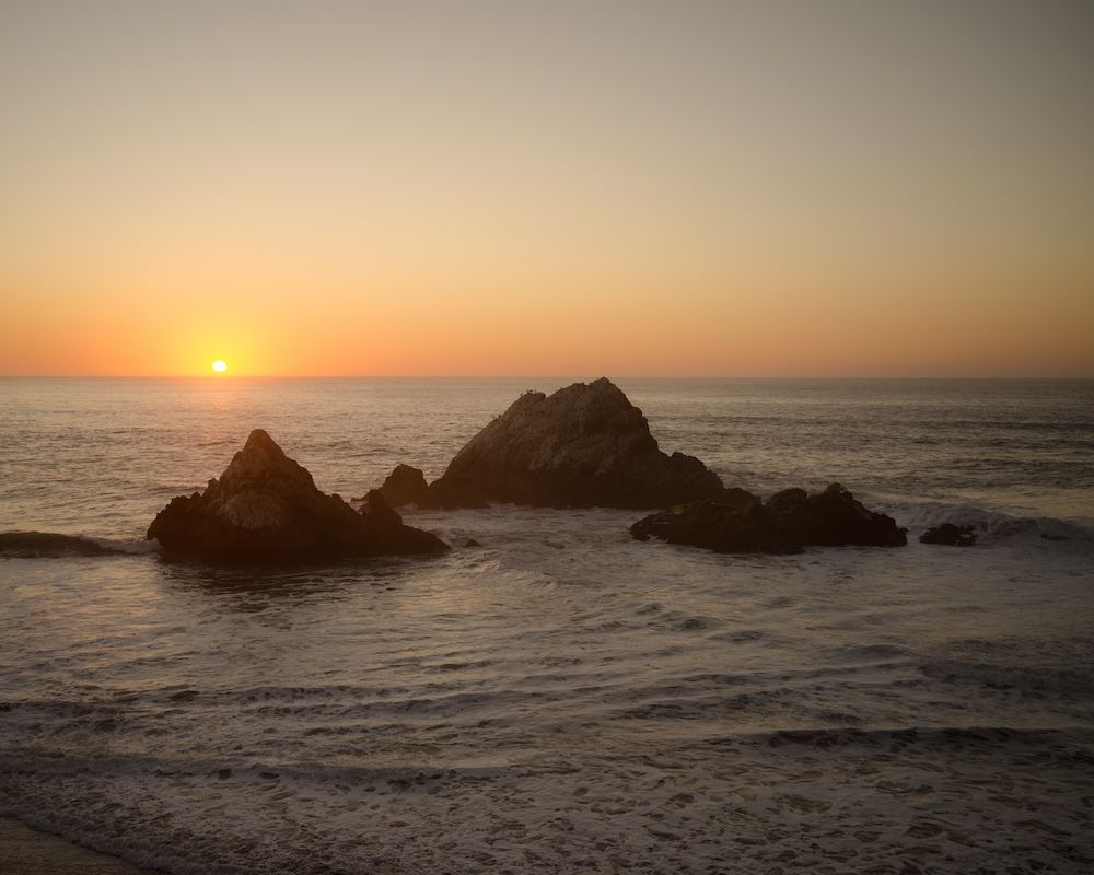 Dramatic sunset over rocky ocean outcrops with golden light reflecting on the water.