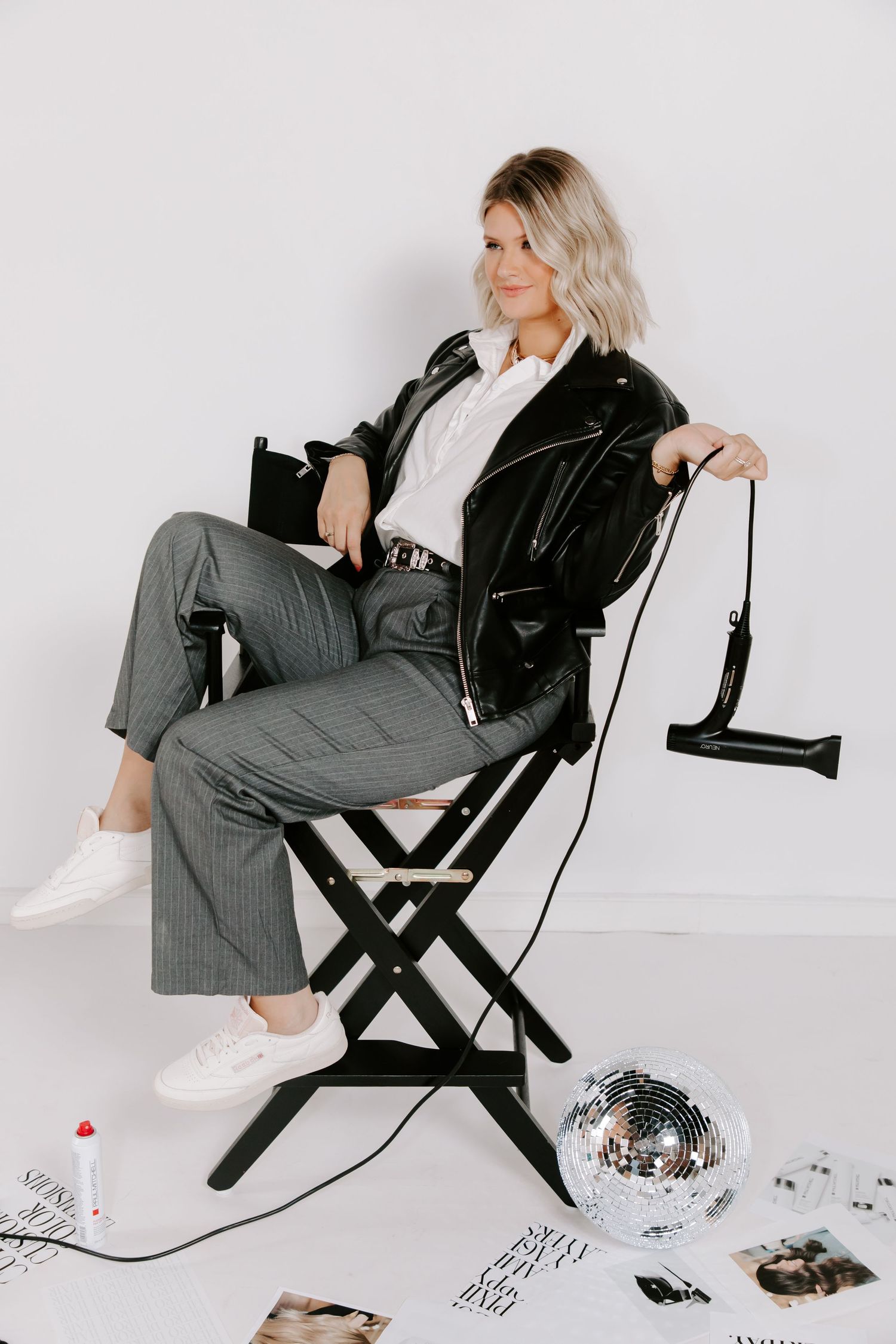 Woman in leather jacket and grey pants poses on director's chair with disco ball.