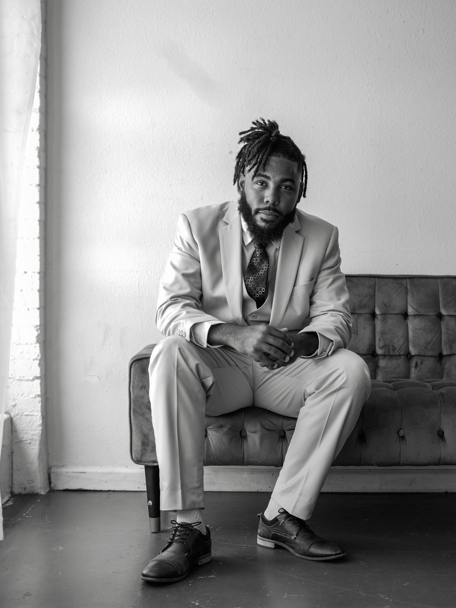 A person in a light gray suit sits thoughtfully on a dark tufted couch in a minimalist black and white setting.