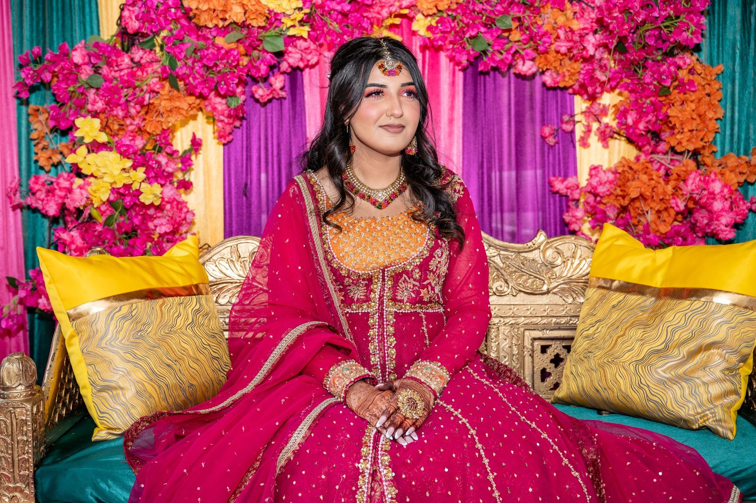 Bride in red and orange lehenga posing before vibrant floral backdrop.