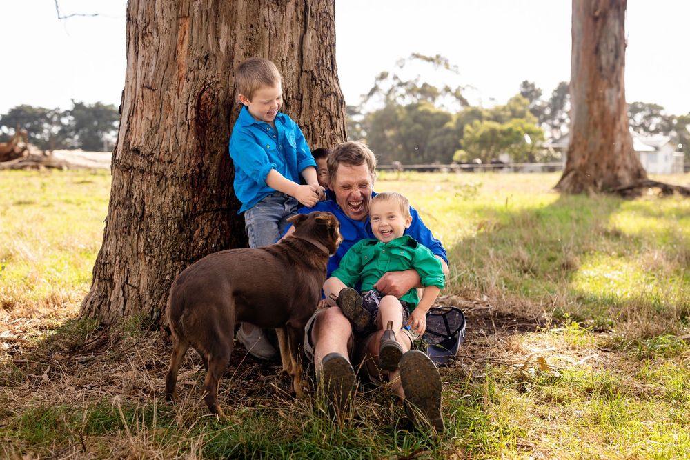 Father and two young boys laughing with a chocolate Labrador under a tree in a sunny rural Australian setting.