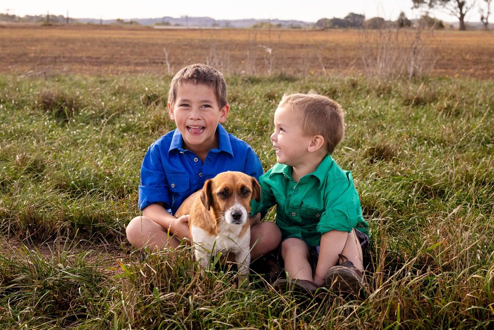 Two young boys in blue and green shirts sit smiling with a beagle puppy in a grassy field.