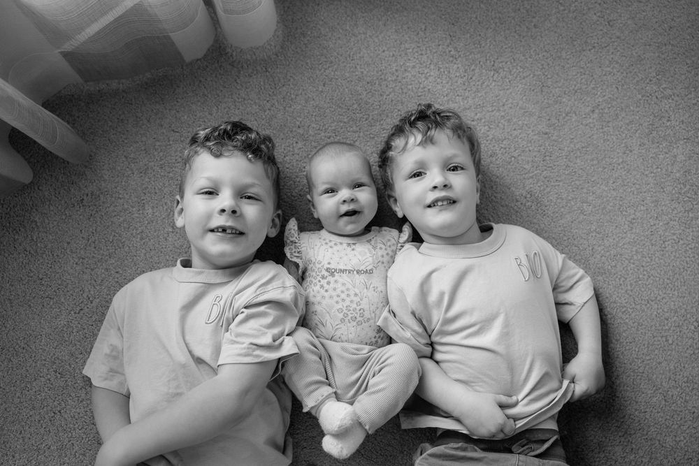 Black and white photo of three young siblings lying on carpet, two older boys flanking a smiling baby girl.