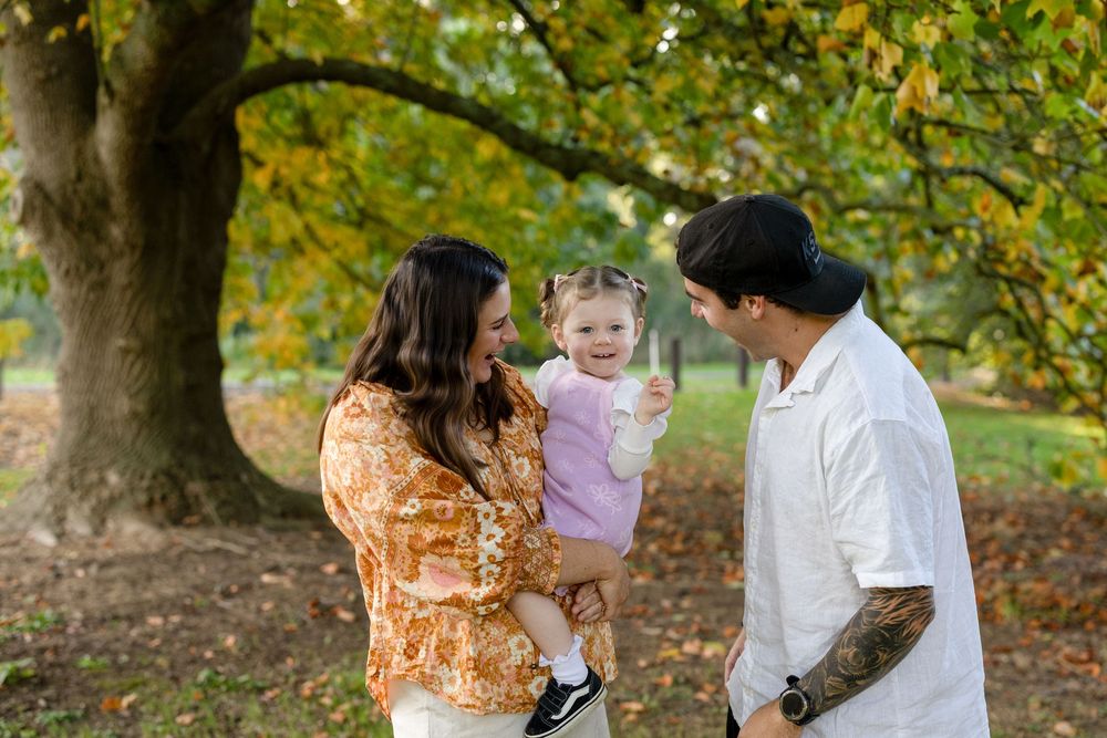 Family of three enjoying autumn park, mother holding toddler girl, father smiling, colorful fall foliage background.