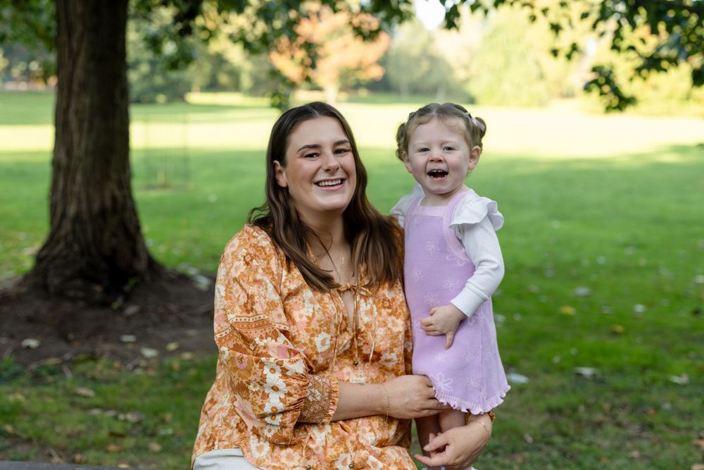 A smiling woman in a floral dress holds a laughing toddler girl in a pink dress outdoors in a green park.