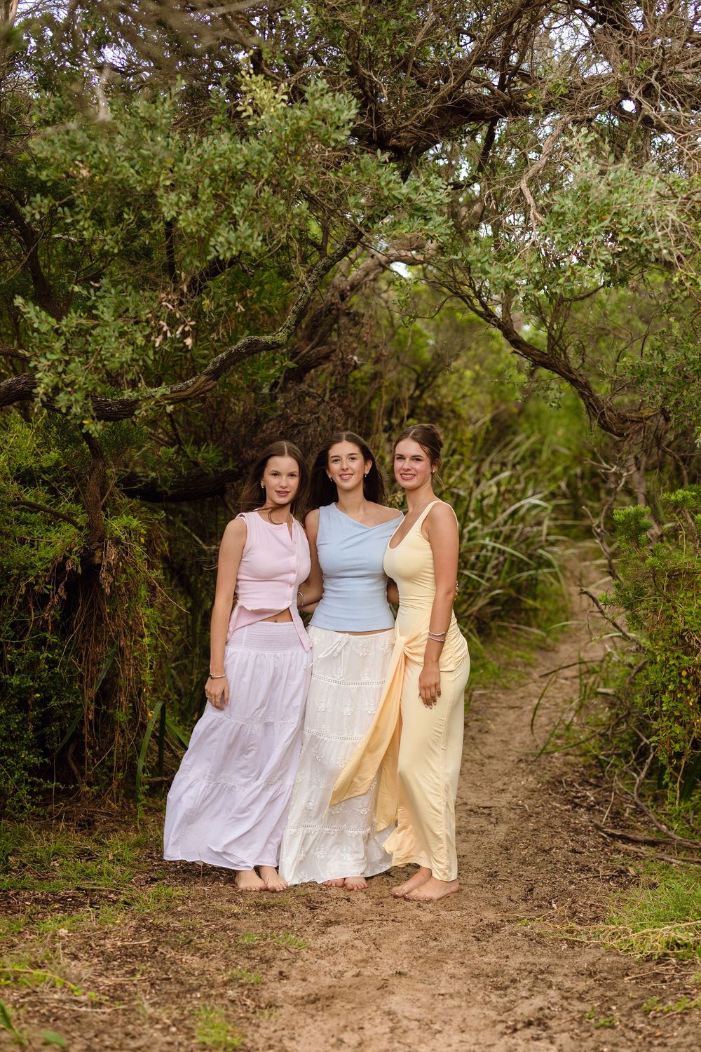 Three women in pastel outfits pose on a forest path beneath arching green trees.