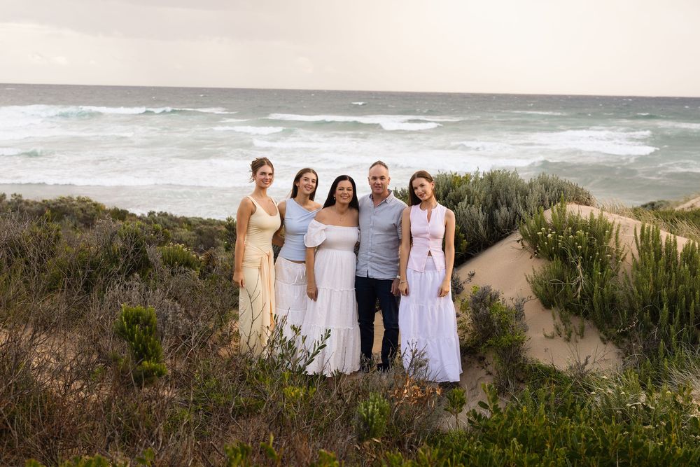 Family of five posing together on sandy coastal dunes with ocean waves in the background at sunset.