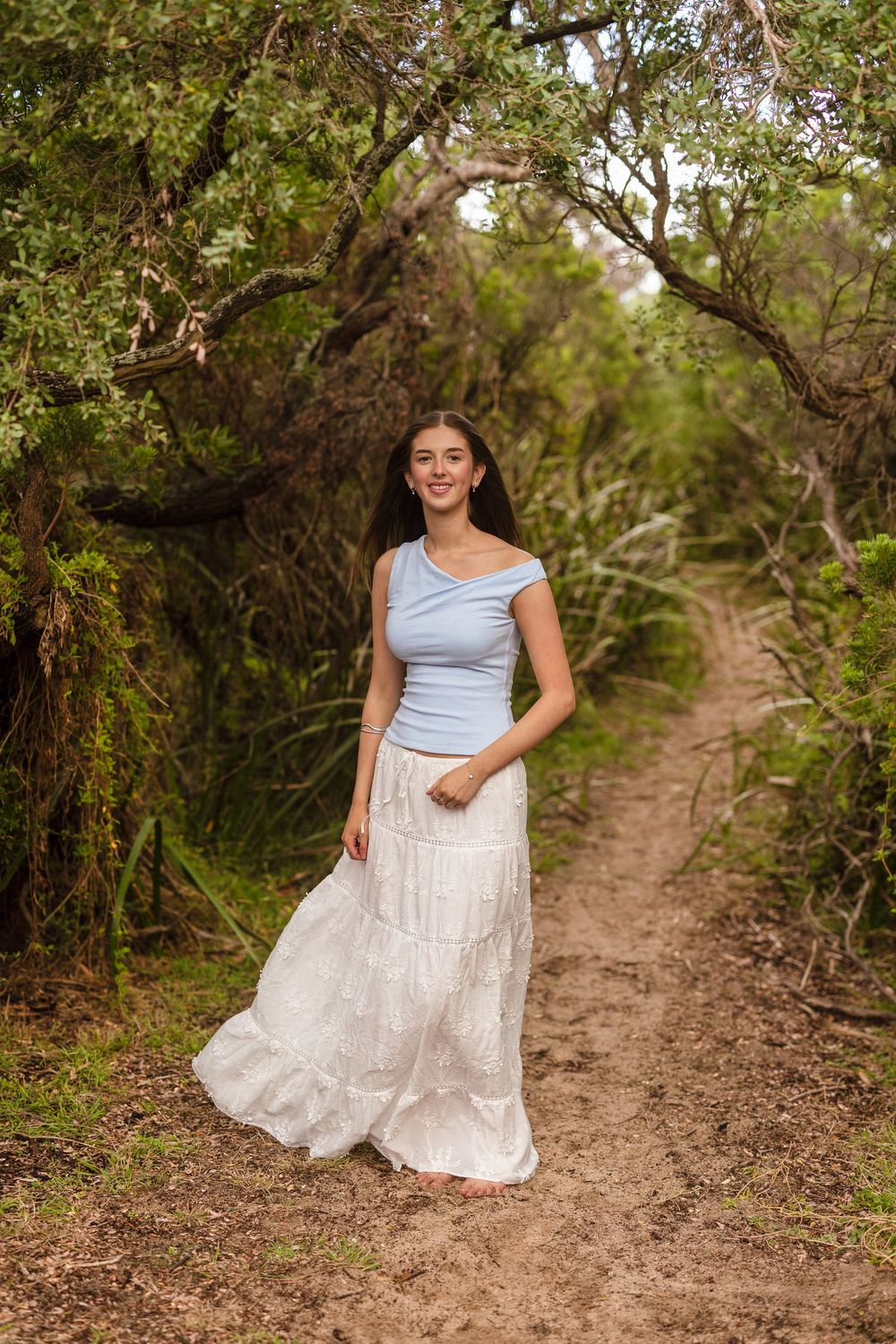 Young woman in blue top and white maxi skirt smiling on a forest trail surrounded by lush greenery.