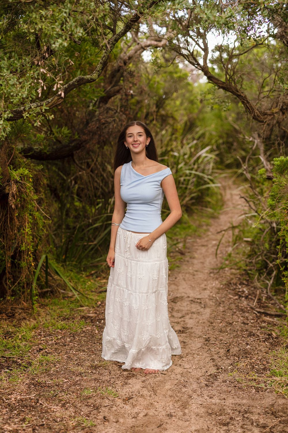 Smiling young woman wearing a light blue top and flowing white skirt standing on a scenic woodland path.