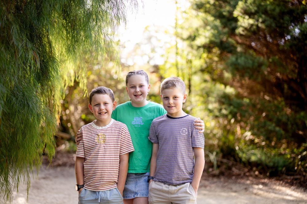 Three smiling children posing together on a sunny garden path surrounded by lush green trees.