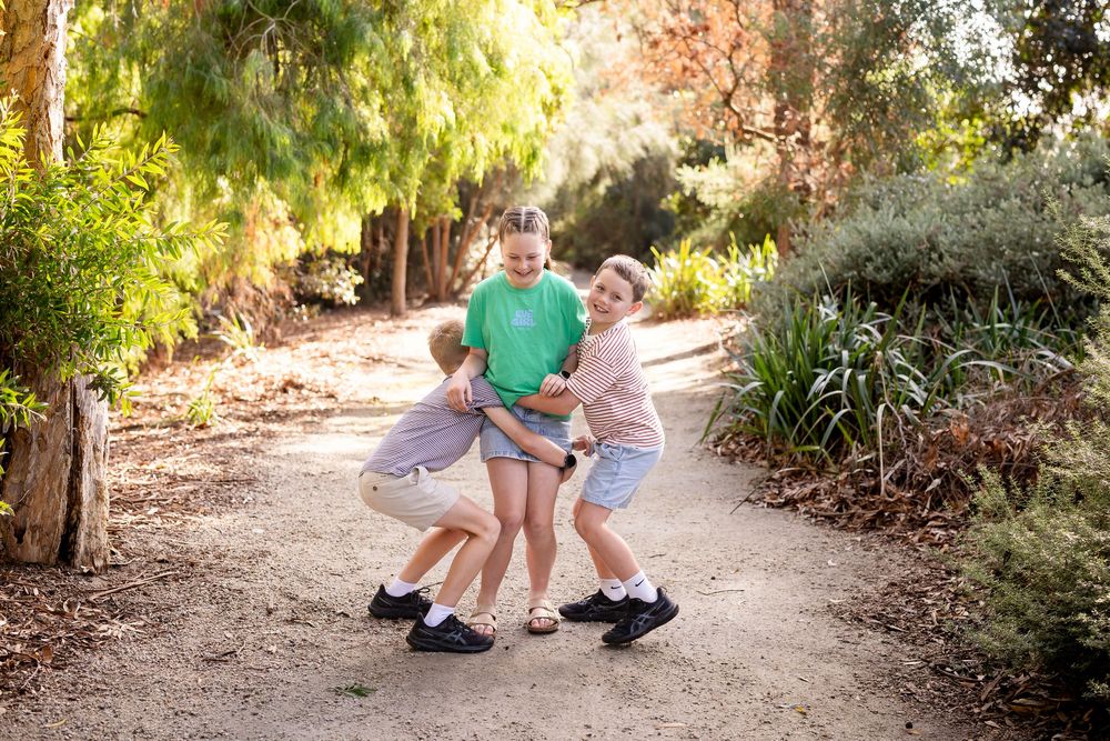 Three children playing and hugging on a dirt path surrounded by lush green trees in a sunny outdoor park.