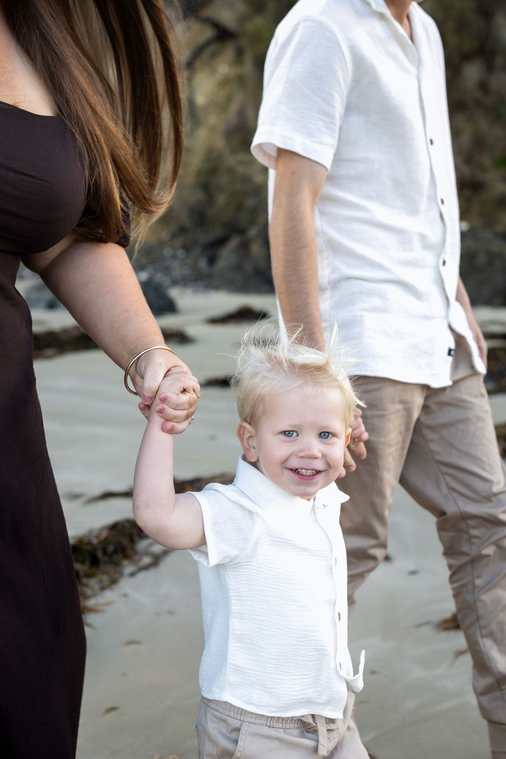 A happy toddler holds parents hands on a sandy beach, wearing a white shirt and khaki pants.