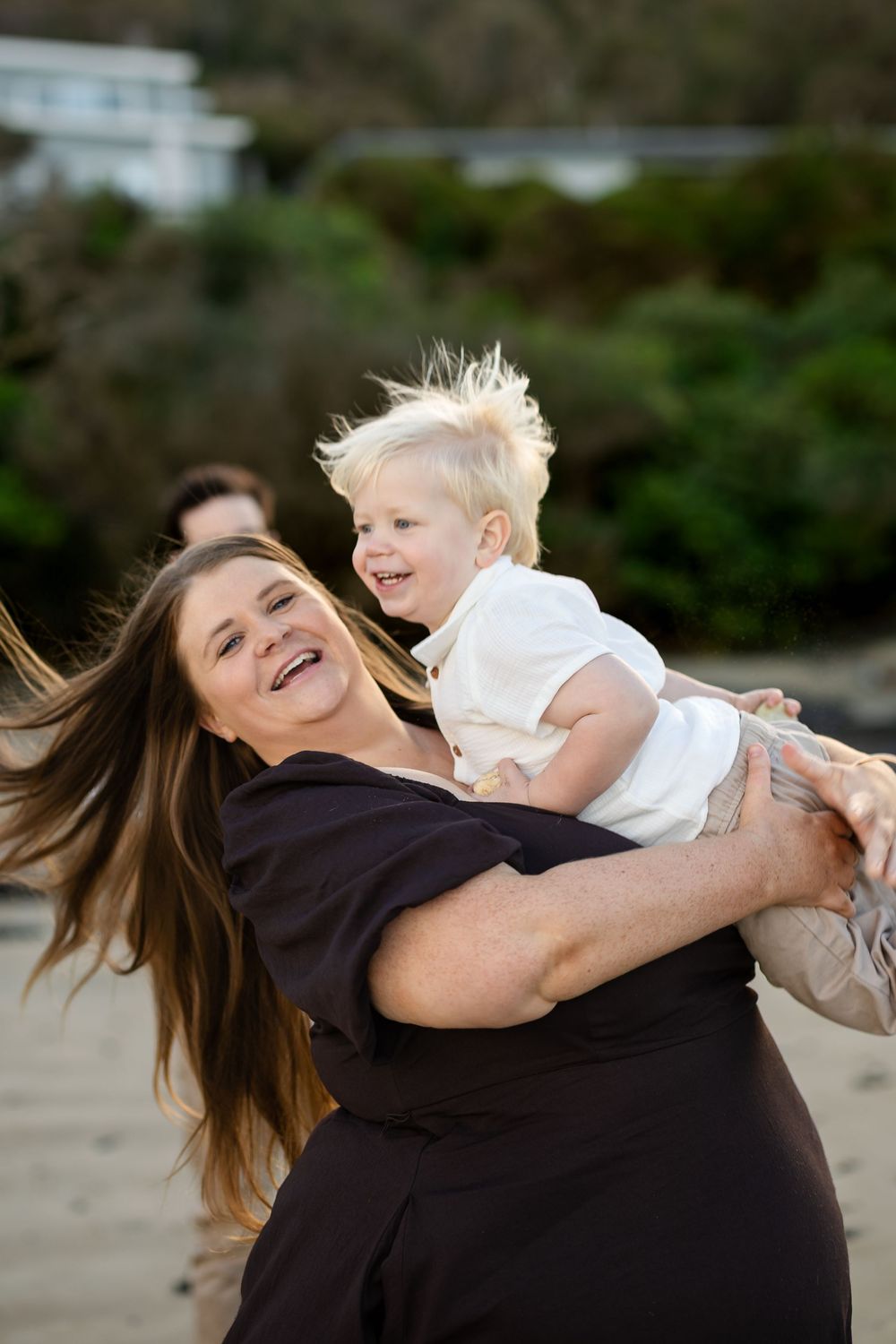 A joyful woman in black dress spins a laughing blonde toddler on a beach, both smiling with wind-blown hair.