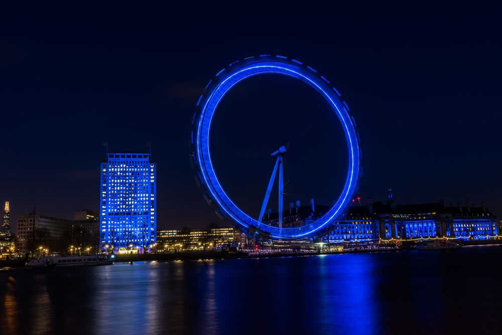 The London Eye and South Bank buildings illuminated in bright blue lights against the dark night sky.