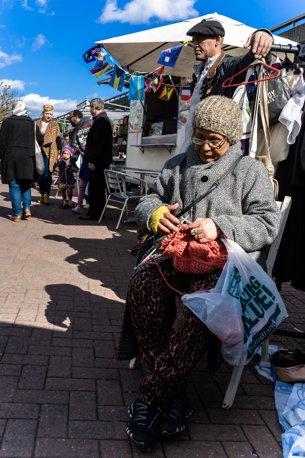 Elderly person in a gray sweater holding shopping bags while sitting in an outdoor market area.