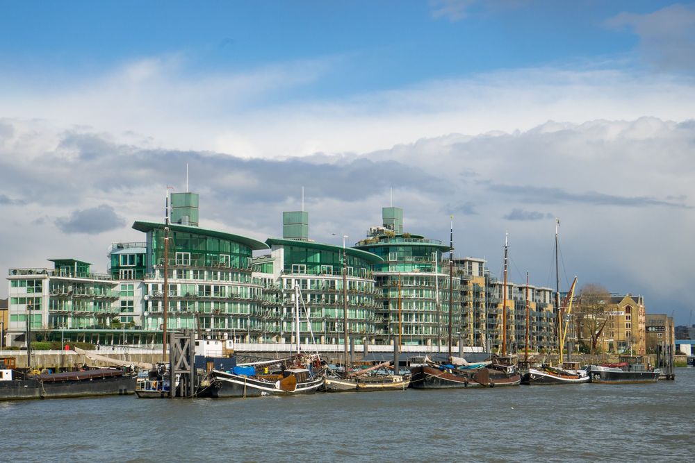 Modern waterfront development with green glass buildings and harbor views along shoreline on cloudy day.