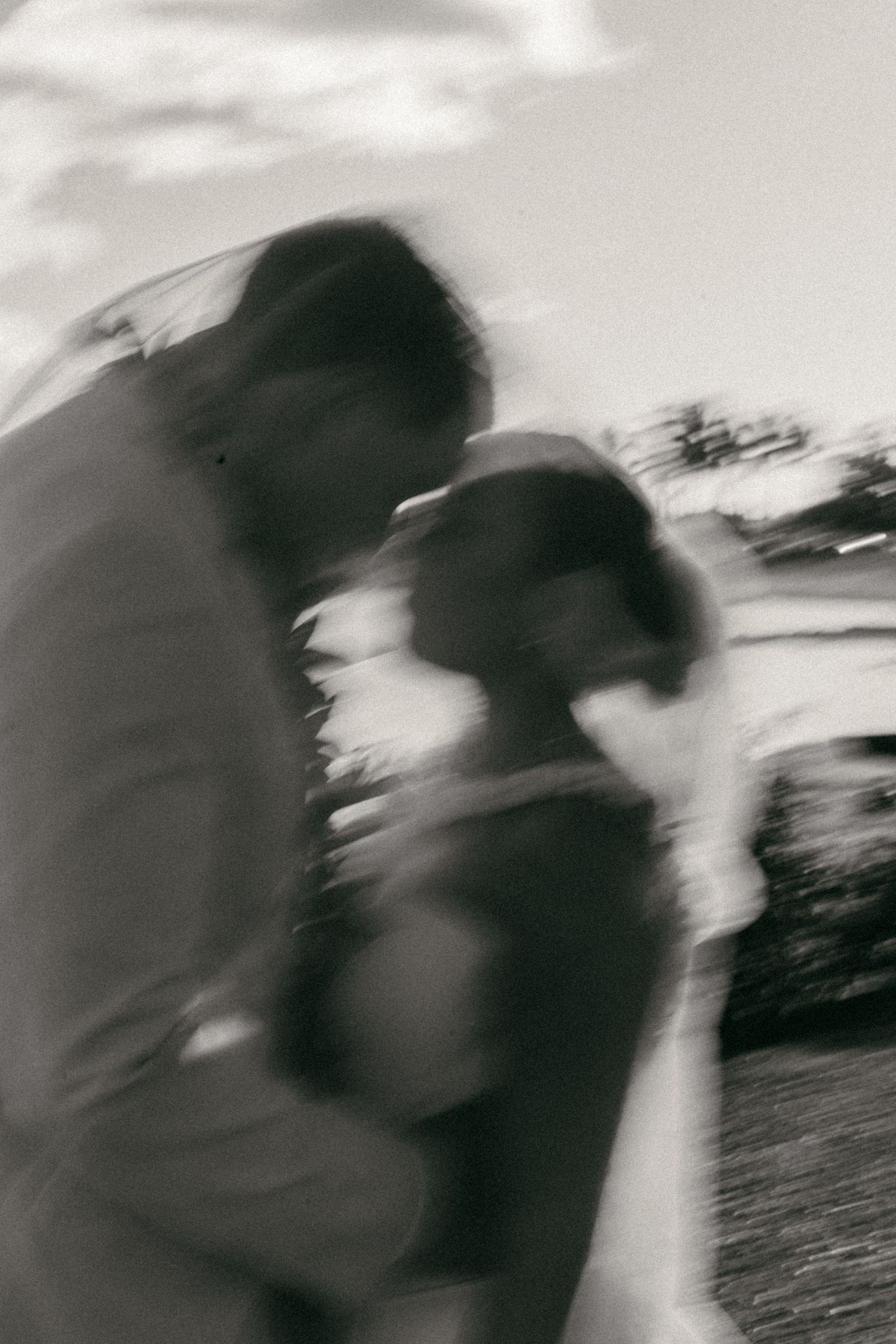 An artistic black and white motion-blurred photograph showing two figures in an intimate embrace outdoors.