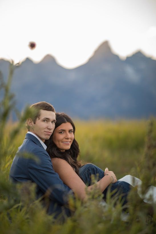 A romantic couple sits together in a field at sunset with mountains visible in the background.
