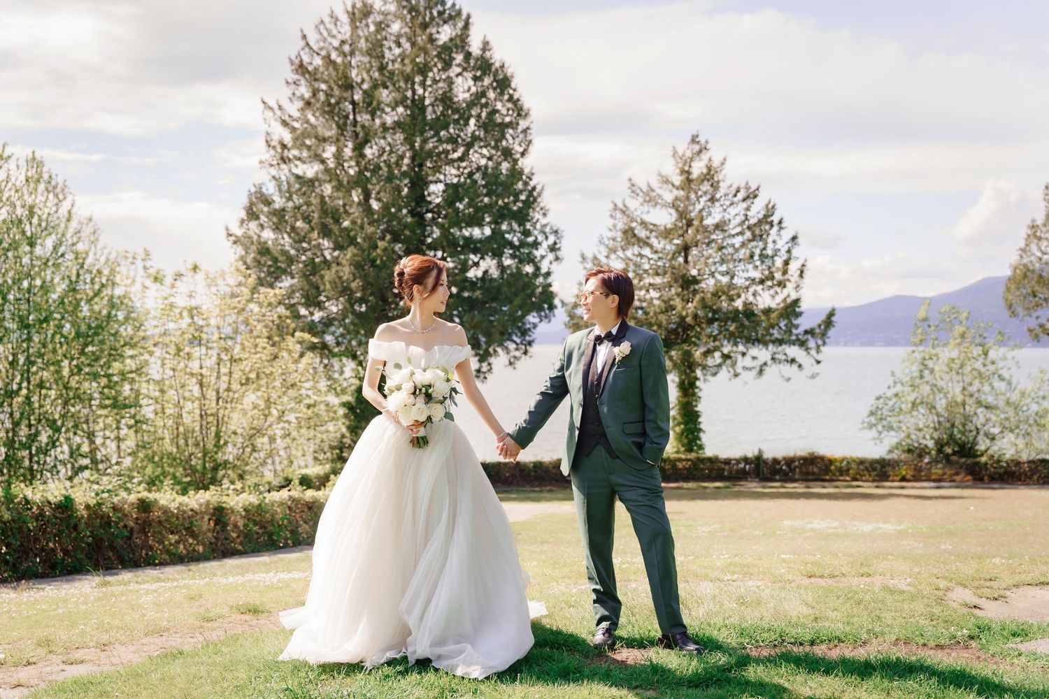 A wedding couple stands on a scenic overlook with evergreen trees and water views in the background.